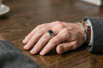 Main d'homme portant une chevalière onyx noire associée à un bracelet argenté et une montre à bracelet cuir, posée sur une table en chêne sombre